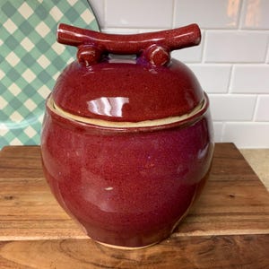 May include: A handcrafted, round, burgundy-colored ceramic jar with a lid. The lid features a handle-like design. The jar sits on a wooden surface, with a green and white checkered plate in the background. The jar is likely used for storage.