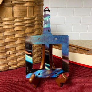 Puede incluir: Una placa de cubierta de interruptor doble con una silueta de faro y un diseño de aurora boreal azul y púrpura. La placa está en un soporte de madera.