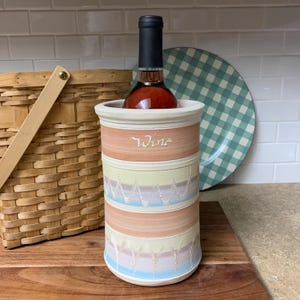May include: A ceramic wine chiller with a bottle of wine inside. The chiller is cylindrical with horizontal bands of color: peach, yellow, and blue. The word "Wine" is written on the peach band. A woven basket and a checkered plate are in the background.