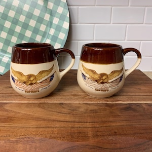 May include: Two brown and cream-colored ceramic mugs with a patriotic eagle design. The mugs have a rounded shape with a brown rim and handle. The eagle is depicted with outstretched wings, and the text "The Spirit of 1776" is printed below.