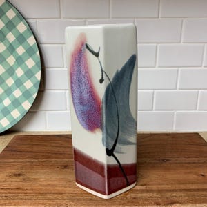 May include: A rectangular white ceramic vase with abstract brushstroke designs in shades of purple, blue, and black. The base of the vase is a deep burgundy color. The vase is displayed on a wooden surface.