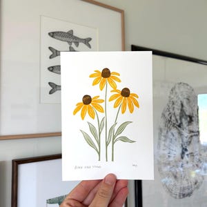 May include: A white rectangular art print featuring a botanical illustration of three Black-Eyed Susan flowers with bright yellow petals, dark brown centres, and sage green stems and leaves. The text "BLACK-EYED SUSANS" is printed in the bottom left corner.
