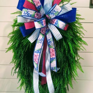 May include: A decorative wreath featuring a lush green base and a vibrant bow. The bow incorporates blue, pink, and white striped ribbons, along with ribbons adorned with a floral hydrangea pattern. The wreath is designed for home decor.
