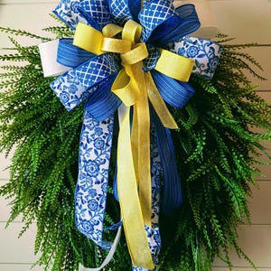 May include: A decorative wreath featuring a lush green base adorned with a large bow. The bow is composed of blue and white patterned ribbons, accented with yellow and gold ribbons. The wreath is suitable for seasonal home decor.