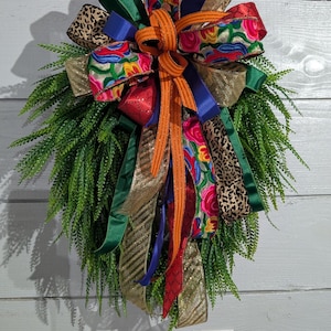 Puede incluir: Una corona decorativa con una vibrante mezcla de colores y texturas. Incluye follaje verde, un gran lazo con cintas estampadas en rojo, azul, dorado y naranja, y cintas con estampado de leopardo. La corona está sobre un fondo de madera blanca.