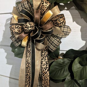 Puede incluir: Una corona decorativa con hojas de magnolia de color verde oscuro y un gran lazo de varias capas. El lazo está hecho de cintas con diversos patrones, incluyendo cuadros negros y beige, estampado de leopardo, negro sólido, oro metálico y marrón.