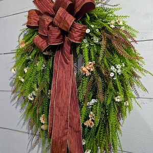 May include: A decorative wreath featuring lush green fern-like foliage, small white and peach-colored flowers, and a large, textured dark red ribbon bow with long, flowing tails that drape down the center of the circular arrangement.
