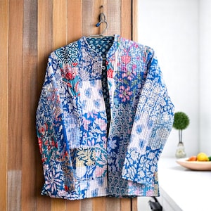 May include: A quilted jacket with a patchwork design. The jacket features a mix of floral and geometric patterns in shades of blue, red, and white. It has a round collar and is hanging on a metal hanger against a wooden door.