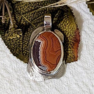May include: A silver pendant featuring a brown and white agate stone. The stone has a swirling pattern of brown, white, and red.