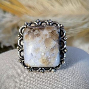 Pode incluir: Um anel quadrado com uma pedra preciosa em uma moldura de filigrana prateada. A pedra apresenta um padr&atilde;o manchado de branco, creme e marrom. O anel est&aacute; sobre uma superf&iacute;cie de cor neutra, com um fundo desfocado.