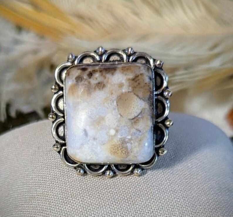 Pode incluir: Um anel quadrado com uma pedra preciosa em uma moldura de filigrana prateada. A pedra apresenta um padr&atilde;o manchado de branco, creme e marrom. O anel est&aacute; sobre uma superf&iacute;cie de cor neutra, com um fundo desfocado.