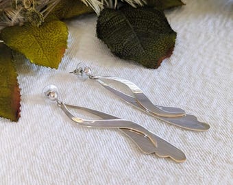 Può includere: Un paio di orecchini pendenti color argento. Ogni orecchino presenta un design fluido e stratificato con un perno arrotondato e un bordo decorativo smerlato. Gli orecchini sono esposti su una superficie bianca.