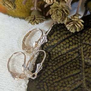 May include: Close-up of three silver-colored earring clasps. The clasps are in various positions, showcasing their design and functionality. The background features a textured green and white surface with floral accents, highlighting the jewelry's delicate details.