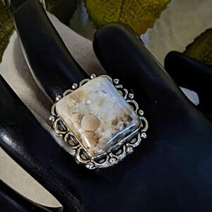 Pode incluir: Anel de prata com uma pedra quadrada de cor clara com padr&otilde;es castanhos e bege. A pedra &eacute; colocada numa moldura de prata decorativa com uma borda ondulada. Um anel marcante, adequado para uso di&aacute;rio ou ocasi&otilde;es especiais.
