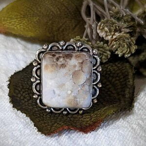 Pode incluir: Anel de prata com uma pedra retangular de cor clara. A pedra tem um padr&atilde;o manchado de branco e castanho. O anel tem uma borda decorativa ondulada.