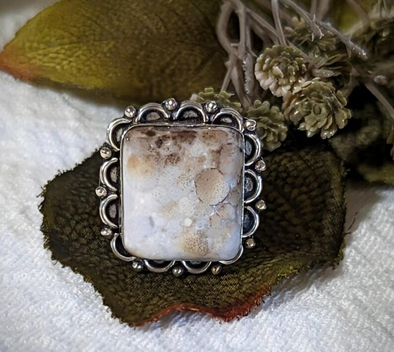 Pode incluir: Anel de prata com uma pedra retangular de cor clara. A pedra tem um padr&atilde;o manchado de branco e castanho. O anel tem uma borda decorativa ondulada.