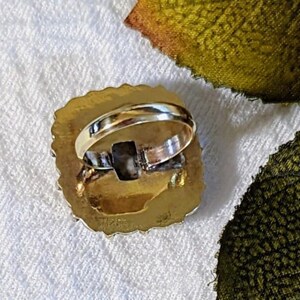 Pode incluir: Um anel dourado com uma pedra quadrada de cor escura embutida em uma moldura oval texturizada. A banda do anel &eacute; de cor dourada polida. O anel est&aacute; sobre uma superf&iacute;cie branca texturizada.