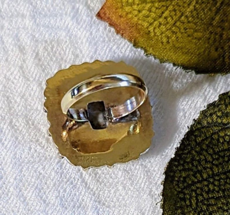 Pode incluir: Um anel dourado com uma pedra quadrada de cor escura embutida em uma moldura oval texturizada. A banda do anel &eacute; de cor dourada polida. O anel est&aacute; sobre uma superf&iacute;cie branca texturizada.