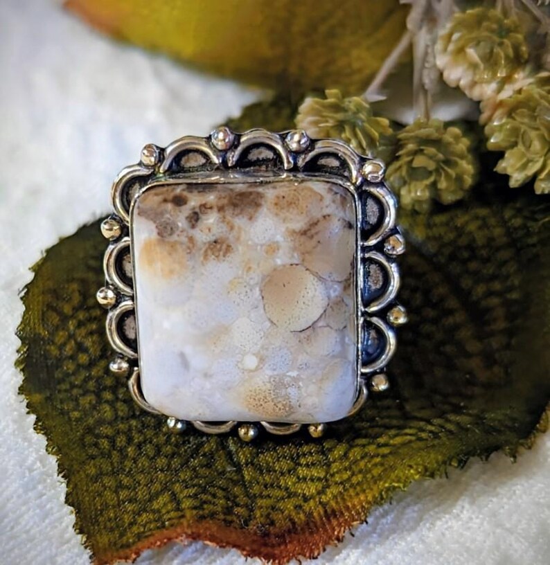 Pode incluir: Um anel prateado com uma pedra retangular grande. A pedra tem um padr&atilde;o mosqueado creme e castanho. O anel tem uma borda decorativa com vieiras. O anel est&aacute; sobre uma folha verde.