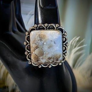 Pode incluir: Anel prateado com uma pedra retangular. A pedra tem um padr&atilde;o mosqueado creme e castanho. O anel tem uma borda decorativa ondulada. O anel &eacute; exibido em uma superf&iacute;cie preta. A pedra tem cerca de 2 cm de comprimento.
