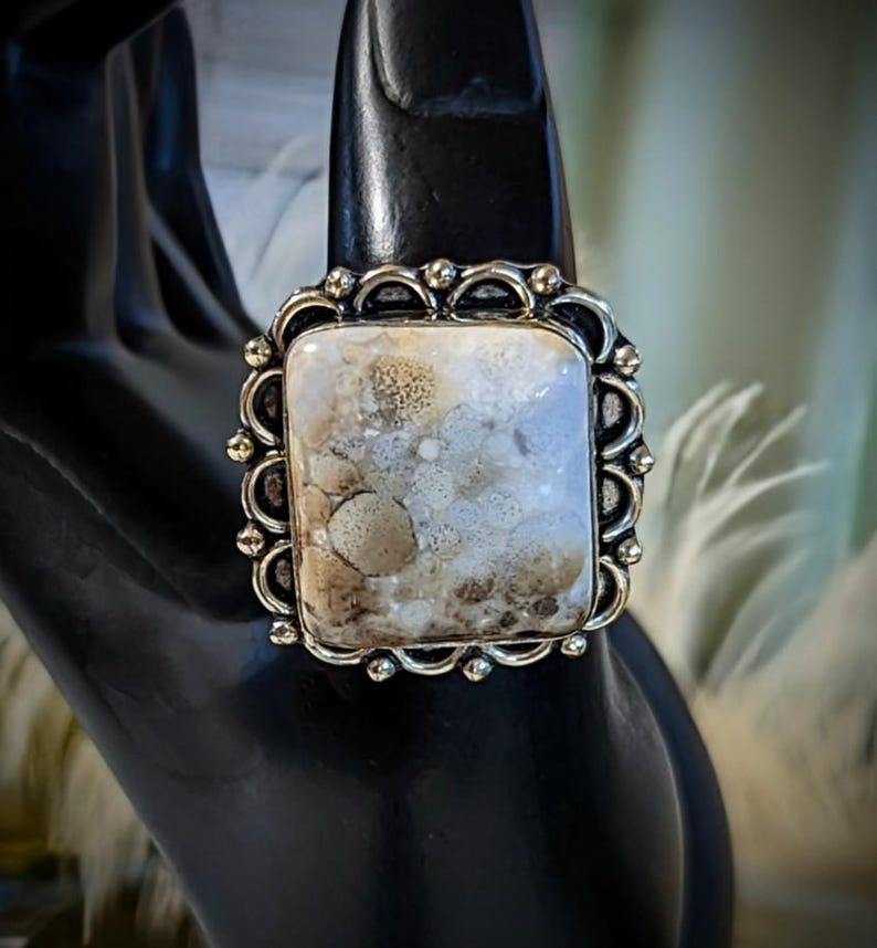 Pode incluir: Anel prateado com uma pedra retangular. A pedra tem um padr&atilde;o mosqueado creme e castanho. O anel tem uma borda decorativa ondulada. O anel &eacute; exibido em uma superf&iacute;cie preta. A pedra tem cerca de 2 cm de comprimento.