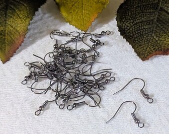 May include: A pile of gunmetal-colored earring hooks with coiled springs and small ball accents. The hooks are scattered on a white surface, with a few individual hooks separated from the main pile. Green and yellow leaves are in the background.