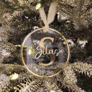 May include: A clear, circular Christmas bauble with a gold trim and the name "Silas" in white script. The letter "S" is gold. It hangs from a cream ribbon against a decorated Christmas tree background, with fairy lights.