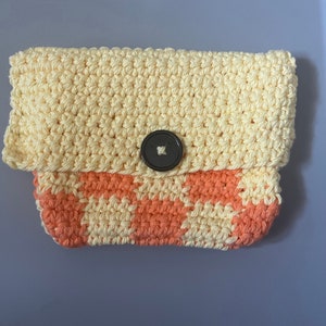 Könnte beinhalten: Eine kleine gehäkelte Clutch mit einem gelben und orangefarbenen Karomuster und einem schwarzen Knopfverschluss.