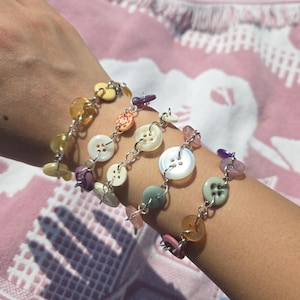 May include: A multi-strand bracelet featuring various colourful plastic buttons in shades of yellow, white, purple, orange, green, and pink, all linked together with silver-toned metal jump rings to create a layered, chain-like accessory design.