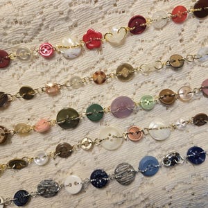 May include: Five strands of decorative chains featuring a variety of buttons in diverse colours, shapes, and sizes. The buttons are linked together with small metal jump rings, creating long, flexible strings of mixed materials and textures.