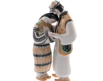 Op de afbeelding: Een keramieken beeldje dat twee figuren in traditionele kledij afbeeldt, die elkaar omarmen. De gedetailleerde kleding heeft patronen in zwart, beige en groen. De man draagt een hoed en de vrouw heeft een bloemenhoofddeksel. Het beeldje staat tegen een witte achtergrond.