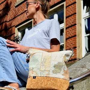 May include: A tote bag with a map design on the top half and a cork base. The bag has a silver zipper and a small leather handle. The map design features shades of green, beige, and brown.