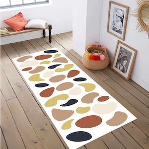 Op de afbeelding: Een rechthoekig wit tapijt met een abstract patroon van organische vormen in verschillende maten, in tinten bruin, beige, geel en marineblauw. Het tapijt ligt op een houten vloer en voegt een moderne touch toe aan de kamer.