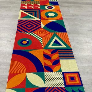 Op de afbeelding: Een lang, rechthoekig tapijt met een geometrisch patroon in tinten rood, oranje, geel, groen, blauw en wit. Het tapijt is gemaakt van verschillende vormen, waaronder cirkels, vierkanten, driehoeken en rechthoeken.