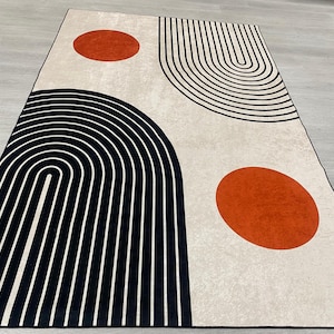 May include: Rectangular rug with a cream background and a modern geometric design. The rug features black curved lines and two orange circles. The design is symmetrical and the rug is placed on a light grey surface.
