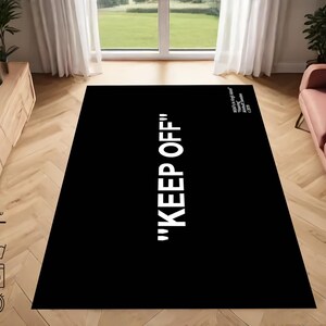 Puede incluir: Alfombra de &aacute;rea negra y blanca con el texto "KEEP OFF" en letras blancas. La alfombra est&aacute; en un ambiente de sala de estar con un sof&aacute; y un centro de entretenimiento de madera.