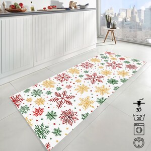 Op de afbeelding: Een rechthoekig wit keukenkleed met een sneeuwvlokkenpatroon in rood, groen en goud. De sneeuwvlokken vari&euml;ren in grootte en zijn verspreid over het kleed. Kleine sterren zijn ook aanwezig.