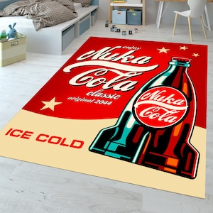 Puede incluir: Una alfombra roja y blanca con una gran imagen de una botella de Nuka Cola. La botella es roja y verde con una etiqueta blanca que dice "Nuka Cola" en una fuente retro. La alfombra también tiene el texto "Enjoy Nuka Cola Classic Original 2044" y "Ice Cold" en blanco.