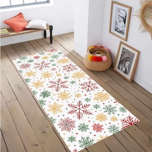 Op de afbeelding: Een rechthoekig wit tapijt met een herhaald patroon van rode, groene en gouden sneeuwvlokken en kleine sterren. Het tapijt ligt op een houten vloer. Een feestelijk, seizoensgebonden woondecoratie-item.