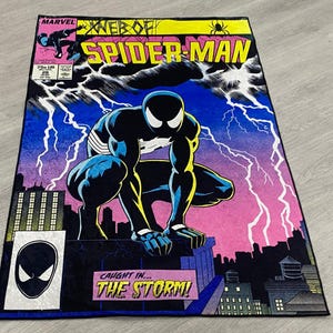 Puede incluir: Una alfombra rectangular con un diseño de portada de cómic de Spider-Man. La alfombra tiene un borde negro y muestra a Spider-Man con un traje en blanco y negro, con rayos y el texto "WEB OF SPIDER-MAN".