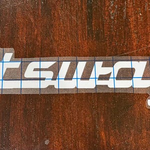 May include: White vinyl decal with the word "Voltswagen" in a stylized font. The decal is on a clear backing with a blue grid pattern, set against a dark wood grain surface.