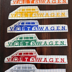 May include: A collection of six vinyl decals featuring a stylized Volkswagen van design. Each decal is a different color: orange, yellow, blue, green, red, and gray. The word "VOLKSWAGEN" is printed below each van.