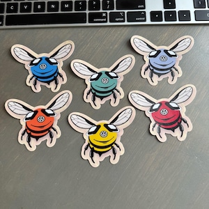 Puede incluir: Seis pegatinas con forma de abeja en varios colores. Cada pegatina presenta una combinación de colores diferente, incluyendo azul, verde, naranja, amarillo, morado y rojo. Las abejas tienen rayas y alas negras, y un logotipo circular en sus cuerpos.