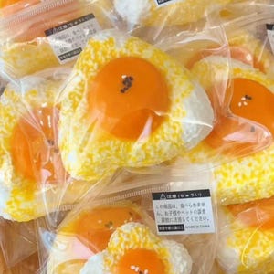 May include: Several packaged artificial fried egg-shaped items are visible. Each has a bright orange yolk and a white and yellow speckled base, resembling rice. They are individually wrapped in clear plastic. The packaging includes Japanese text.