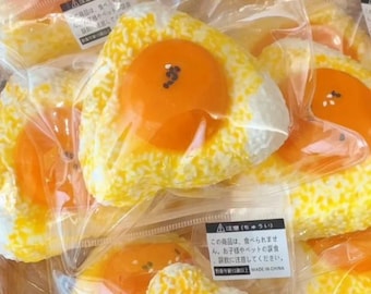 Könnte beinhalten: Nahaufnahme von mehreren verpackten, künstlichen Spiegelei-förmigen Artikeln. Die Eier haben ein leuchtend orangefarbenes Eigelb und eine weiß-gelb gesprenkelte Basis, die Reis ähnelt. Jedes ist einzeln in Klarsichtfolie verpackt.