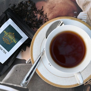 May include: A white teacup with a gold rim and a saucer sits on a magazine. The teacup is filled with dark brown tea. A silver teaspoon rests on the saucer. A black tea bag with the text "FINE INDIA TEA USA PRODUCT OF INDIA" is visible in the background.