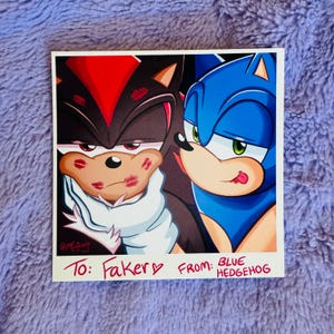 May include: A square print featuring two cartoon hedgehogs, one black and red, the other blue. The black hedgehog has lipstick marks on its face. Text reads "To: Faker" and "From: Blue Hedgehog."