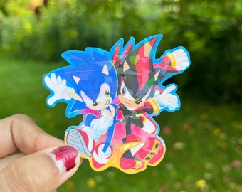 May include: A sticker featuring Sonic the Hedgehog and Shadow the Hedgehog, both in their classic poses. Sonic is blue with white gloves and shoes, while Shadow is black with red accents and gold rings. The sticker has a colorful background with a gradient effect.