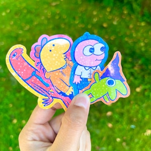 May include: Four colourful cartoon character stickers with glitter accents. The stickers feature a pink character with large eyes, a yellow character with a blue hat, a green character with a blue hat, and a red character with a blue tail.