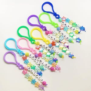 May include: A collection of colorful keychains with names spelled out in white letter beads. Each keychain features a different color clasp, smiley face beads, and star-shaped beads. Names include Olivia, Gavin, Chloe, Jackeli, Ivy, Chad, and Hayes.